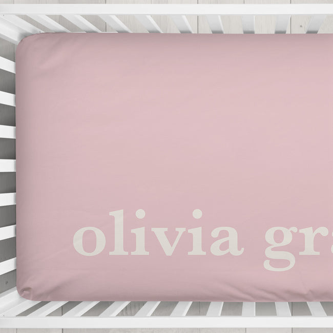 A dusty pink fitted crib sheet with the name 'olivia grace' printed in white lowercase serif letters, shown inside a white wooden crib.