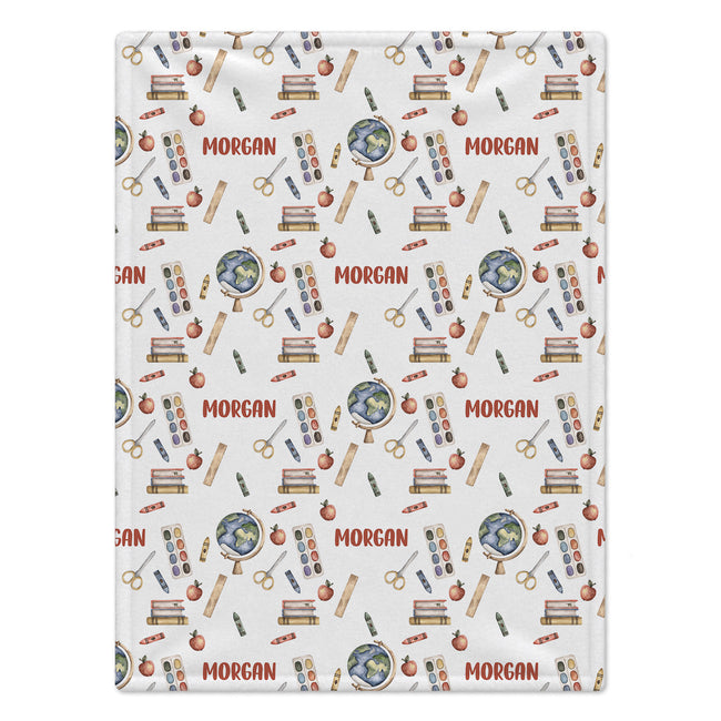 A rectangular white fabric item, likely a baby blanket or towel, featuring a repeating school-themed pattern with books, globes, rulers, pencils, and apples. The name 'MORGAN' is printed repeatedly in red across the design.