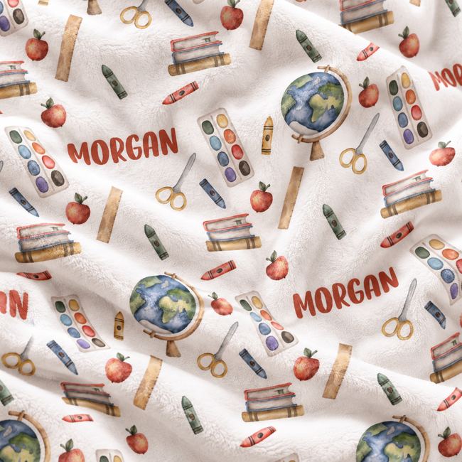 A close-up of a white fleece blanket with a school-themed pattern featuring books, apples, globes, crayons, scissors, paint palettes, and the name 'MORGAN' printed in red.