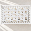 A top-down view of a white crib mattress fitted with a printed sheet featuring cute cartoon cows, clouds, and the text 'KAI' in a warm neutral palette, set inside a white wooden crib.