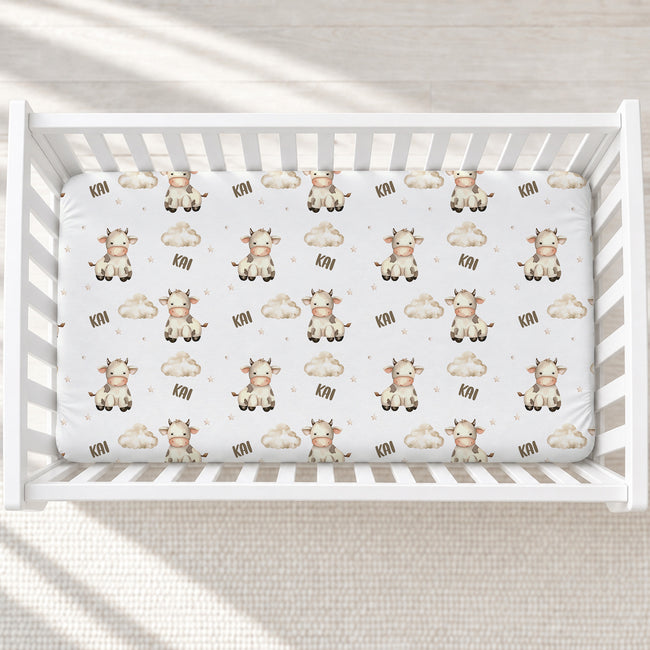 A top-down view of a white crib mattress fitted with a printed sheet featuring cute cartoon cows, clouds, and the text 'KAI' in a warm neutral palette, set inside a white wooden crib.