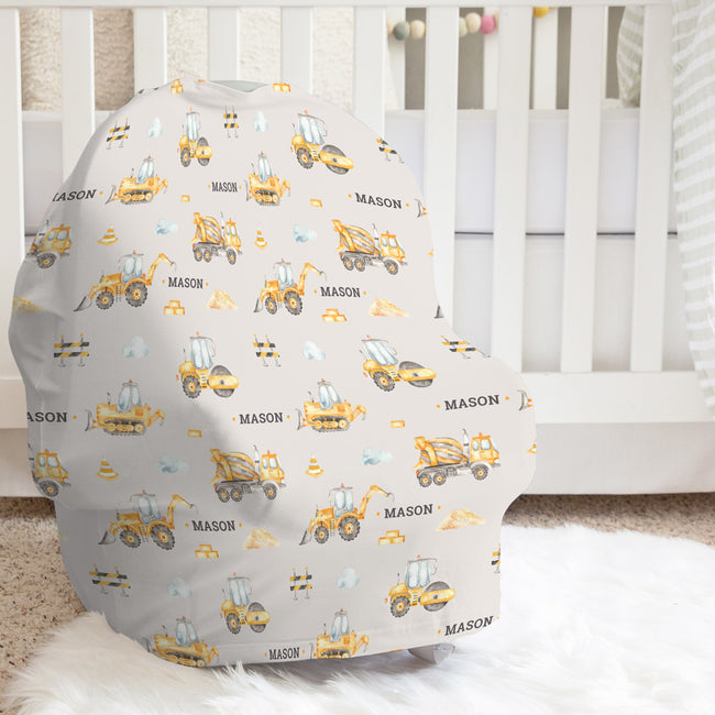 A baby car seat cover with a construction-themed print featuring yellow excavators, blue cranes, and the name 'MASON' on a light gray background, styled in a nursery setting in front of a white crib.