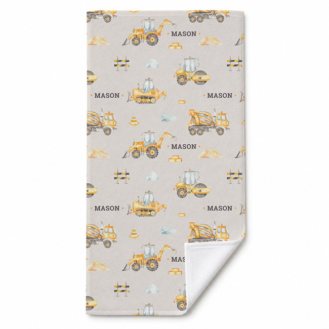 A personalized beach towel with a light gray background and a repeating pattern of yellow construction vehicles, including excavators, bulldozers, and a cement mixer, along with the name 'MASON' printed in black. The bottom corner is folded over to show the white reverse side.