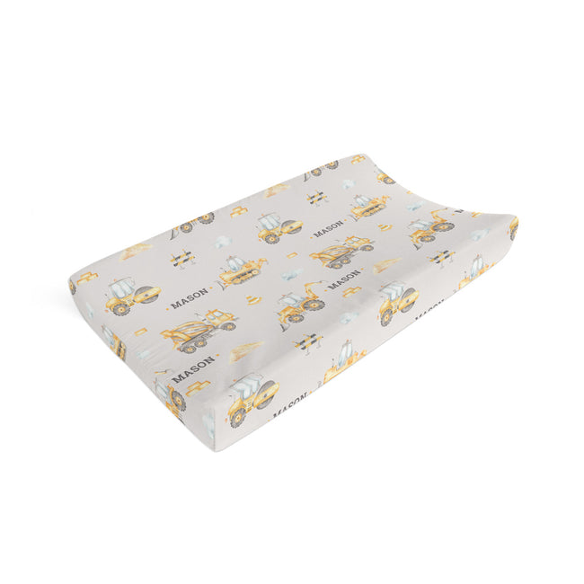 A contoured baby changing pad cover featuring a yellow construction vehicle and sailboat pattern on a light gray background, with the name 'MASON' printed repeatedly.