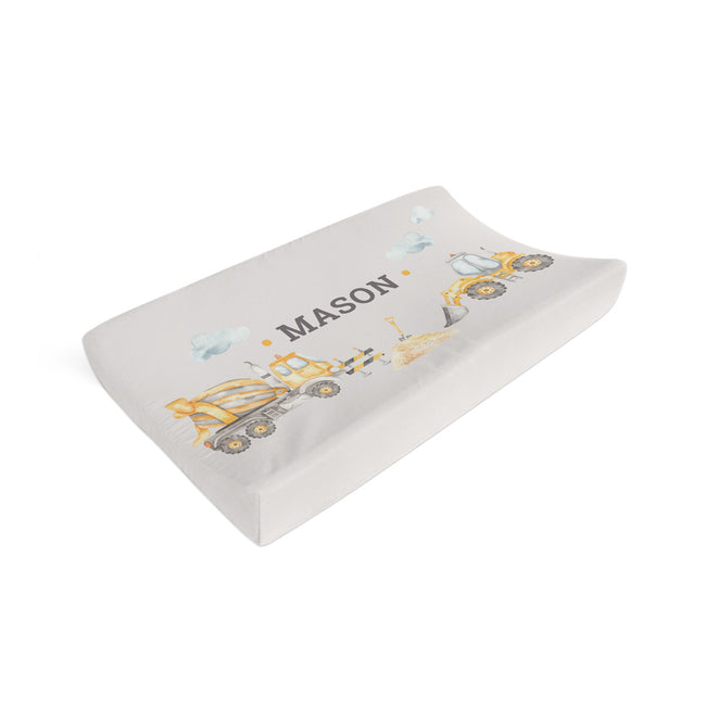 A contoured baby changing pad cover in a light gray fabric with a printed design of yellow construction vehicles and the name 'MASON' in dark gray, shown on a plain white background.