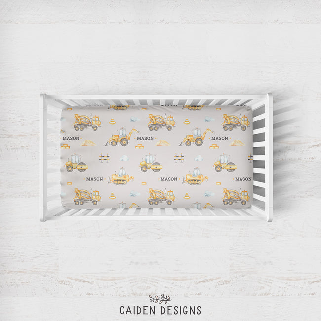 A top-down view of a white wooden crib with a fitted sheet featuring a construction-themed print with yellow excavators, bulldozers, and the name 'MASON' repeated. The crib is set against a light, rustic white wood wall background.