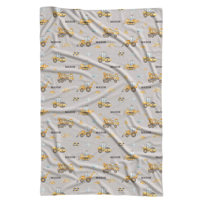 A swaddle blanket with a light gray background and an all-over repeating pattern of yellow construction vehicles and the word 'MASON' in black.