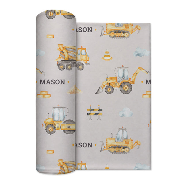 A rolled swaddle blanket with a construction-themed pattern featuring yellow construction vehicles, traffic cones, clouds, and the name 'MASON' on a light gray background.