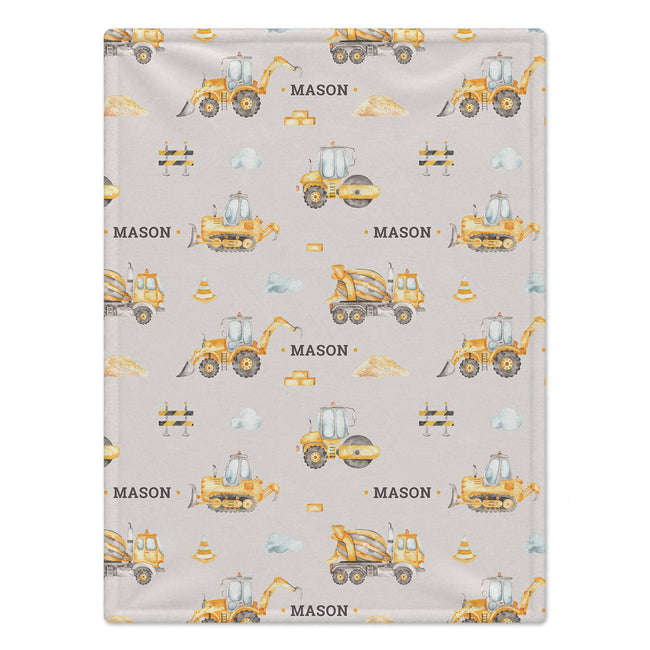 A rectangular baby blanket with a light gray background and a repeating construction-themed pattern featuring yellow and orange excavators, bulldozers, road rollers, traffic cones, and the name 'MASON' printed in black.