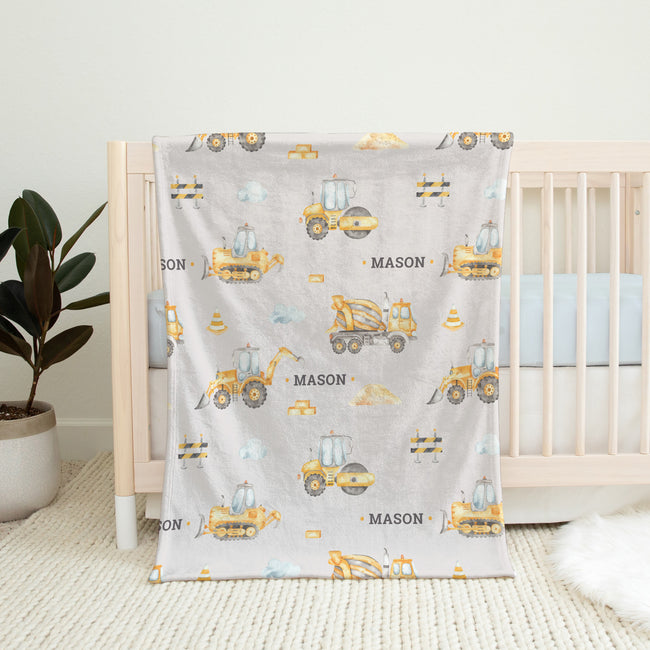 A light gray baby blanket with a construction vehicle pattern is draped over a light wood crib in a nursery setting.