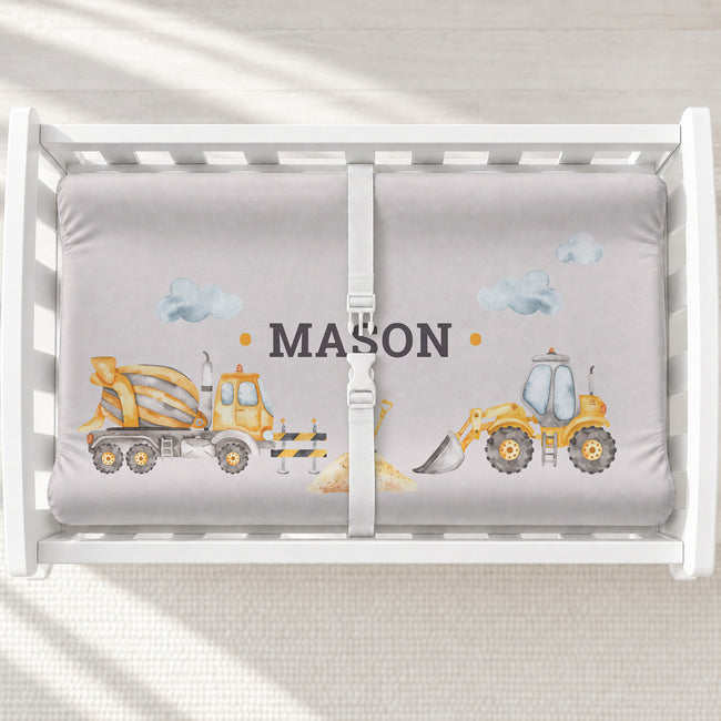 A white changing table with a personalized changing pad cover featuring a light gray background with yellow construction vehicles and the name 'MASON' in the center.