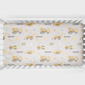 A crib sheet featuring a repeating pattern of yellow construction vehicles and the name 'MASON' on a light beige background, shown inside a white crib.