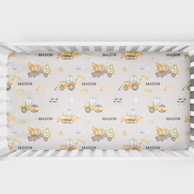 A crib sheet featuring a repeating pattern of yellow construction vehicles and the name 'MASON' on a light beige background, shown inside a white crib.