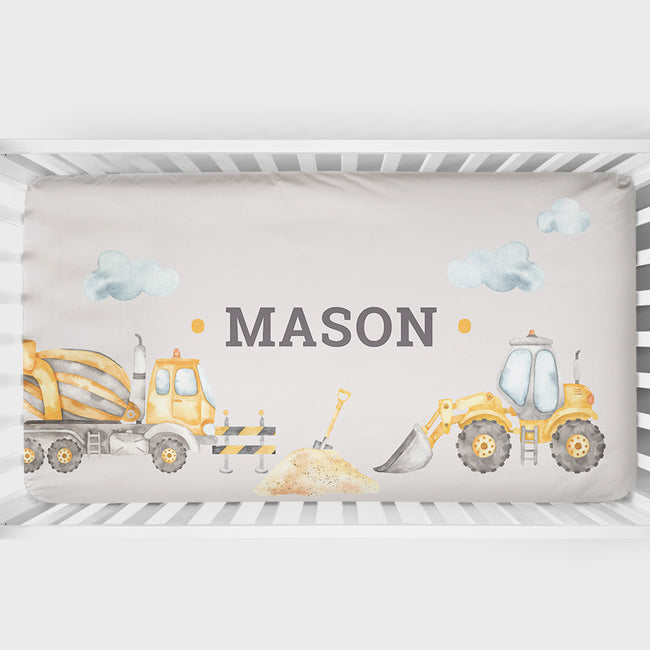 Personalized baby crib bedding featuring a construction-themed design with a cement mixer, excavator, bulldozer, clouds, and the name 'MASON' in gray text on a light beige background.