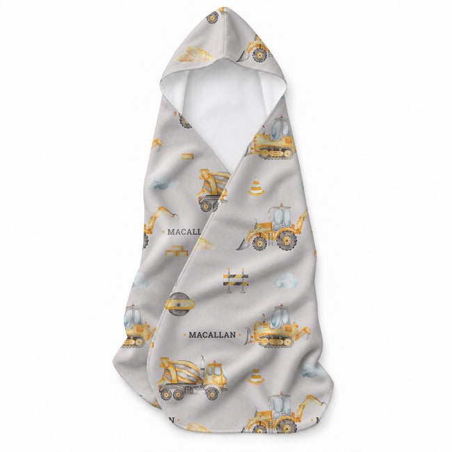Baby hooded towel featuring yellow construction vehicles, traffic cones, bricks, blue clouds, and the name Macallan in gray lettering.