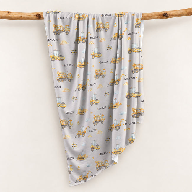 Personalized swaddle blanket hanging on a wooden rail with watercolor construction trucks, excavators, cement mixers and custom name Mason.