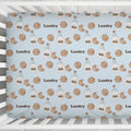A crib sheet with a repeating pattern of chocolate chip cookies, milk bottles, and the word 'Landry' on a light blue background, shown hanging in a white crib.