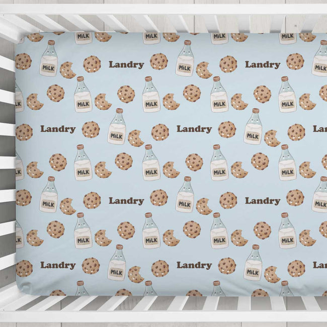 A crib sheet with a repeating pattern of chocolate chip cookies, milk bottles, and the word 'Landry' on a light blue background, shown hanging in a white crib.