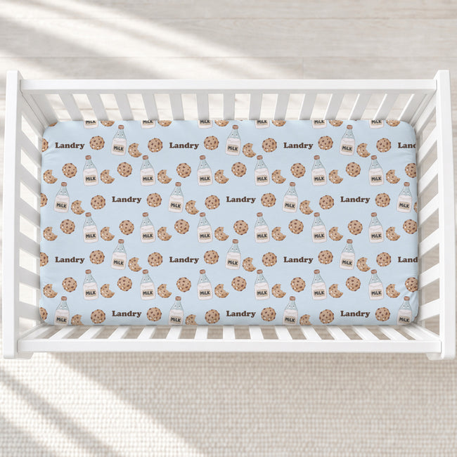 A baby crib with white slats, fitted with a light blue mattress cover printed with a repeating pattern of milk bottles and cookies with the brand name 'Landry' and 'MILK' written on them.