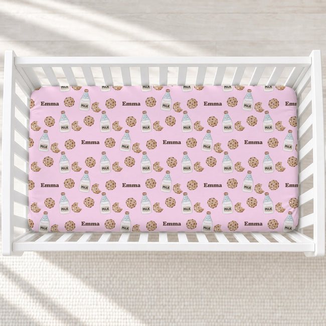 A pink baby crib mattress fitted sheet with a repeating pattern of chocolate chip cookies, milk bottles, and the name 'Emma' in brown, shown inside a white wooden crib.