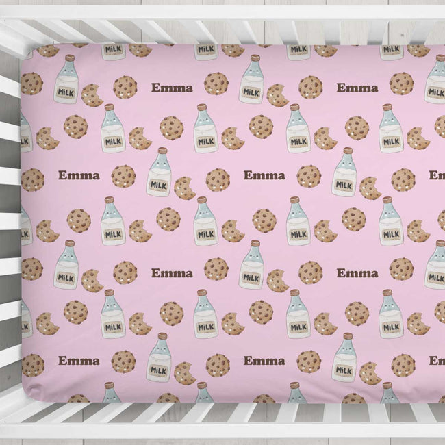 A pink crib fitted sheet with a repeating pattern of chocolate chip cookies, milk bottles, and the name 'Emma' in brown text, shown inside a white wooden crib.
