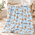 A light blue blanket with a repeating pattern of chocolate chip cookies, milk bottles, and the word 'Landry' is draped over a woven basket in a bright, sunlit room.