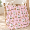 A pink blanket with a repeating pattern of chocolate chip cookies and milk bottles, personalized with the name 'Emma', draped over a woven basket in a bright nursery setting.