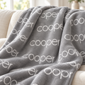 A plush gray throw blanket with a repeating white 'cooper' name pattern, draped over a light beige sofa in a bright indoor setting.