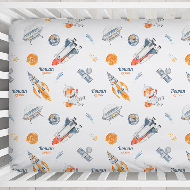A fitted crib sheet with a colorful space-themed print featuring rockets, UFOs, planets, stars, and the personalized name 'Rowan' in blue script, shown inside a white wooden crib.