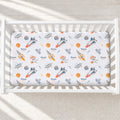 A white crib mattress fitted sheet with a colorful space-themed print featuring rockets, astronauts, planets, and the name 'Noonan' printed repeatedly, shown inside a white wooden crib.