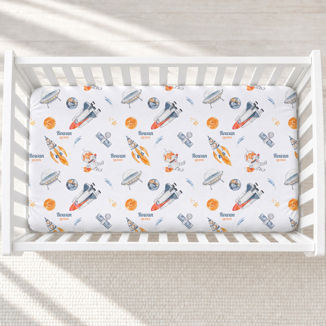 A white crib mattress fitted sheet with a colorful space-themed print featuring rockets, astronauts, planets, and the name 'Noonan' printed repeatedly, shown inside a white wooden crib.