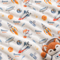 Close-up of a soft fleece blanket with an astronaut-themed print, featuring rockets, UFOs, stars, and the name 'Rowan QUINN' repeated across the cream fabric. A plush orange fox astronaut is visible at the bottom right corner.
