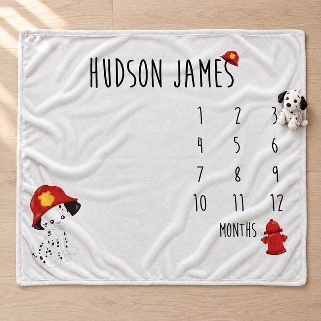 A personalized white baby blanket laid flat on a light wood floor, printed with the name 'HUDSON JAMES' at the top, a 12-month countdown grid, and firefighter-themed illustrations including a Dalmatian puppy in a red helmet and a red fire hydrant.