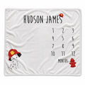 A personalized white blanket featuring a black calendar grid for 12 months, the name 'HUDSON JAMES' at the top, a Dalmatian puppy illustration wearing a red firefighter helmet in the lower left, and a red fire hydrant near the bottom right.