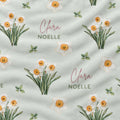 A close-up of a fabric surface featuring a repeating watercolor floral print of narcissus in white, yellow, and green on a pale sage background, with the words 'Clara NOELLE' printed in pink and green script.