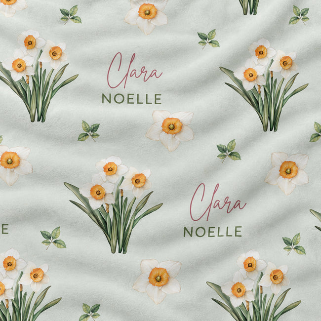 A close-up of a fabric surface featuring a repeating watercolor floral print of narcissus in white, yellow, and green on a pale sage background, with the words 'Clara NOELLE' printed in pink and green script.