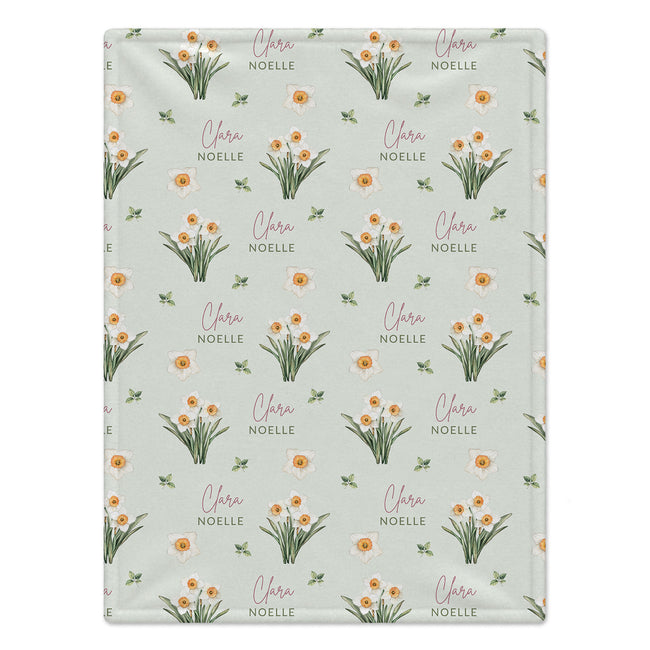 Pale sage blanket with white narcissus flowers, golden-orange centers, green leaves, and the name Clara Noelle in rose and green.