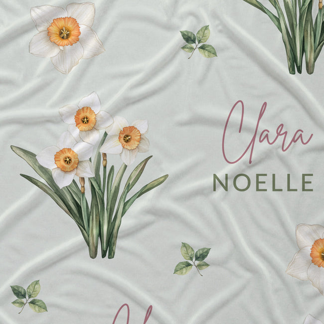 Pale sage fabric with white narcissus flowers, scattered green leaves, and the name Clara Noelle repeated in rose and green.