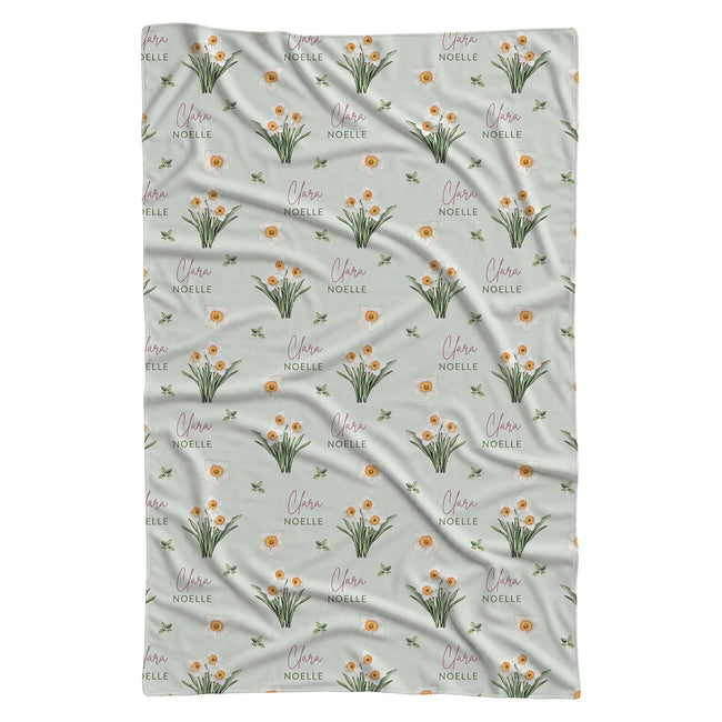 Pale sage swaddle blanket with white narcissus flowers, scattered green leaves, and the name Clara Noelle repeated in rose and green.