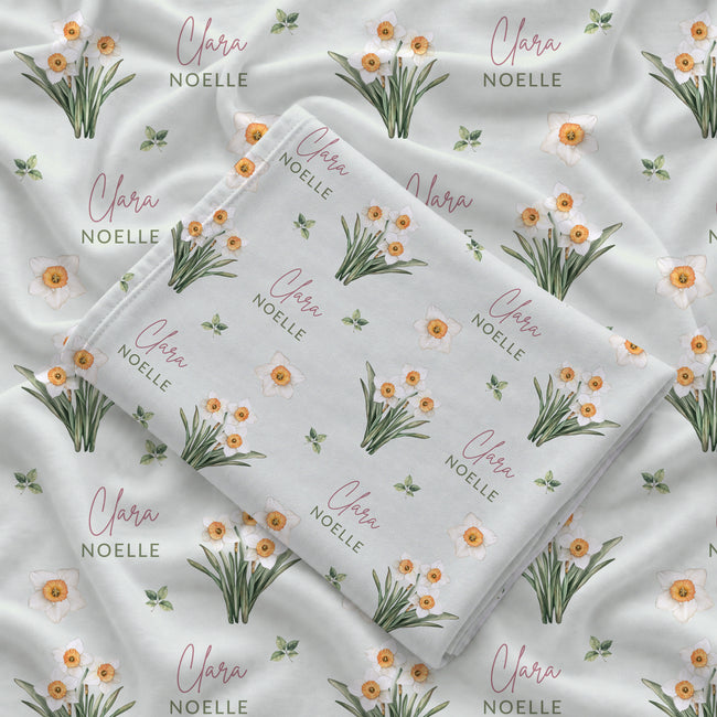 Pale sage swaddle blanket with white narcissus flowers, scattered green leaves, and the name Clara Noelle repeated in rose and green.