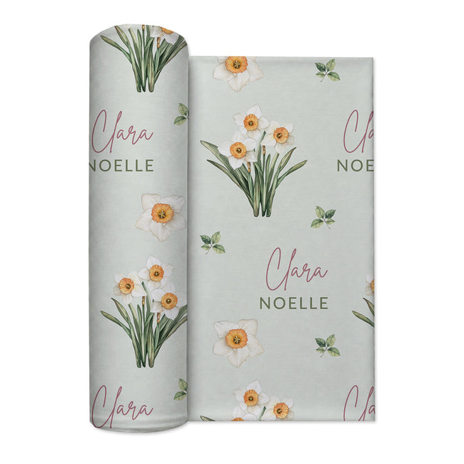 Pale sage swaddle blanket with white narcissus flowers, scattered green leaves, and the name Clara Noelle repeated in rose and green.