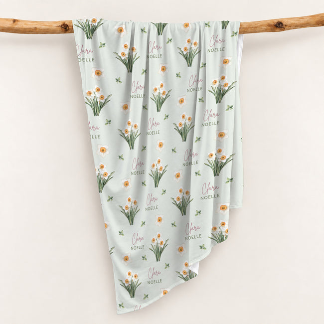 Pale sage swaddle blanket with white narcissus flowers, scattered green leaves, and the name Clara Noelle repeated in rose and green.