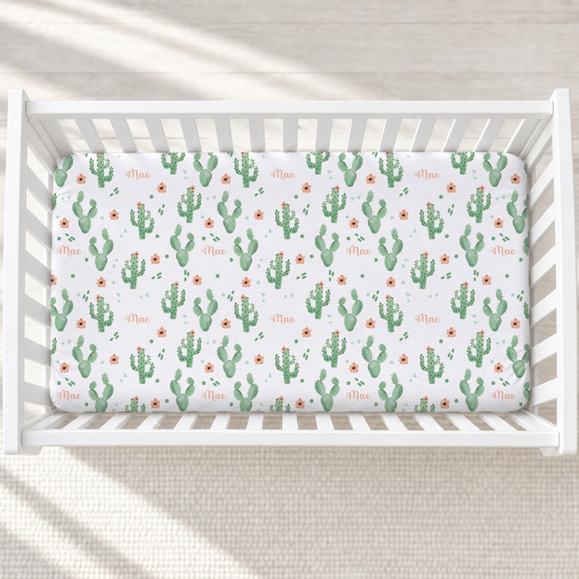 A white wooden crib with a fitted sheet featuring a green cactus and small orange flower pattern, with the name 'Mae' printed in orange script.