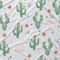 A close-up of a white fabric with a repeating pattern of green watercolor cacti, small peach flowers, green dots, and the name 'Mae' in peach script.