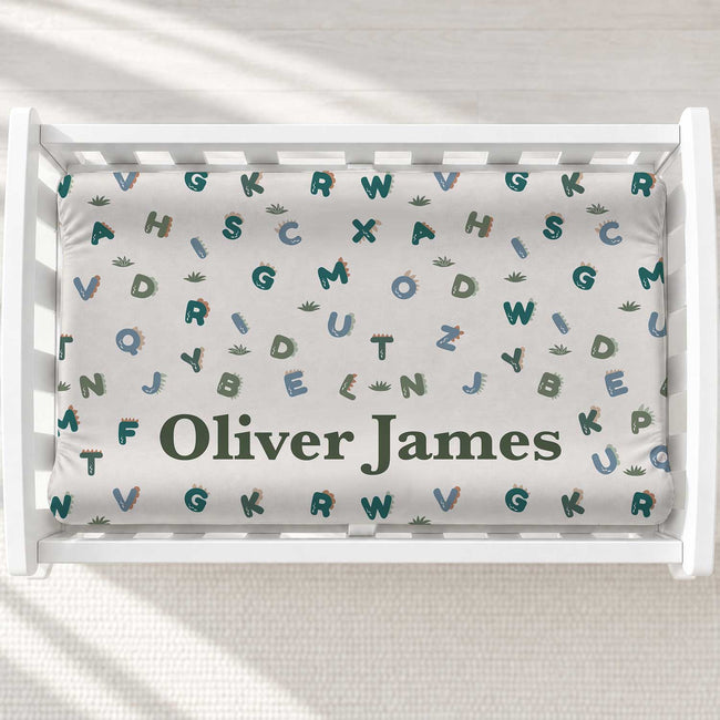 Customized baby changing pad cover featuring an alphabet and cactus pattern in teal, green, and blue on a white background, with the name 'Oliver James' printed in dark green. The bedding is displayed inside a white changing table.