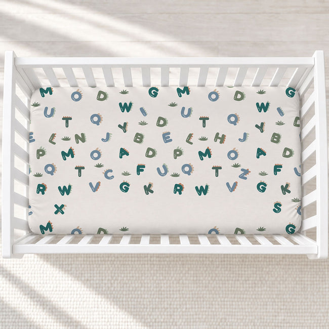 A white crib with a fitted sheet featuring a playful alphabet and dinosaur pattern, photographed from a top-down angle on a light textured floor.