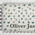 Customized baby crib bedding featuring a cream background with a playful pattern of colorful dinosaur letters and small cactus prints, with the personalized name 'Oliver James' printed in dark green at the bottom.