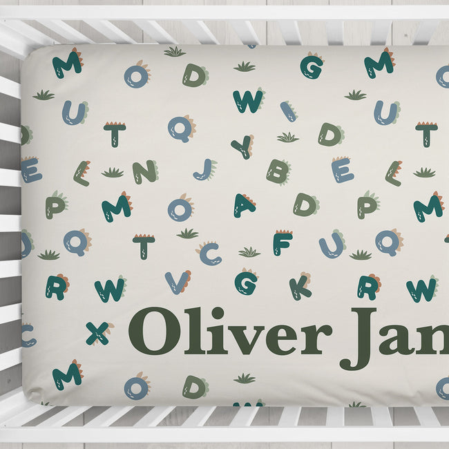 Customized baby crib bedding featuring a cream background with a playful pattern of colorful dinosaur letters and small cactus prints, with the personalized name 'Oliver James' printed in dark green at the bottom.