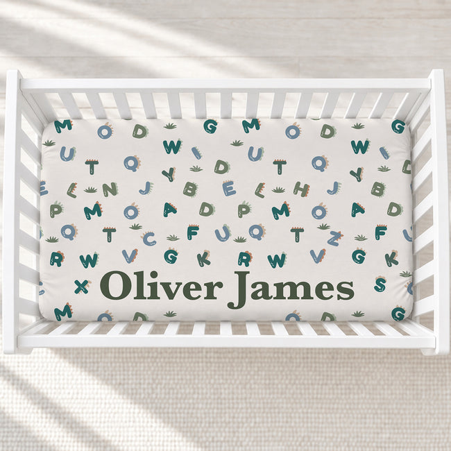 A personalized baby crib mattress inside a white wooden crib, featuring a white background with a pattern of colorful alphabet letters and small dinosaur illustrations, with the name 'Oliver James' printed in dark green at the bottom.