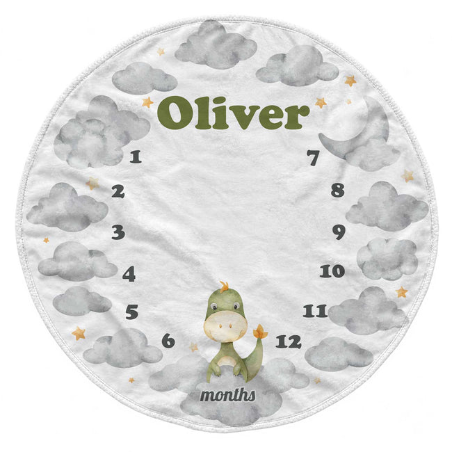 A round baby milestone chart towel or blanket featuring a white background with gray watercolor clouds, a small green dinosaur, the name 'Oliver' in olive green, and numbers 1 through 12 arranged around the circle with the word 'months' at the bottom.